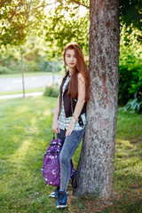 Portrait of beautiful white Caucasian young woman with long red hair, standing with backpack  in park outside at sunset. Autumn fall back-to-school concept. Schoolgirl student.