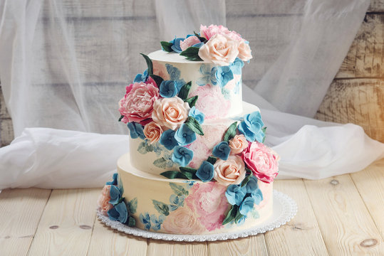 A beautiful home wedding three-tiered cake decorated with pink roses and blue flowers in a rustic style on wooden table