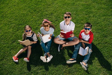 High angle view of cute multiethnic kids in sunglasses reading books while sitting on grass and smiling