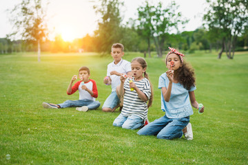 Fototapeta premium Cute multiethnic kids blowing soap bubbles while sitting on green lawn in park