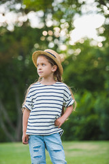 Adorable little girl in straw hat standing with hand on waist and looking away in park