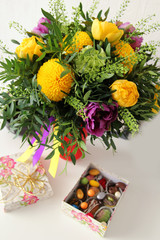 A bouquet of yellow, green and purple flowers on a table with box of chocolate candies.