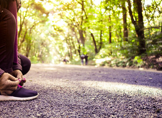 woman with weaved brown wool gloves knee down to do up pink shoelaces in morning light at park, beautiful green tone bokeh in background