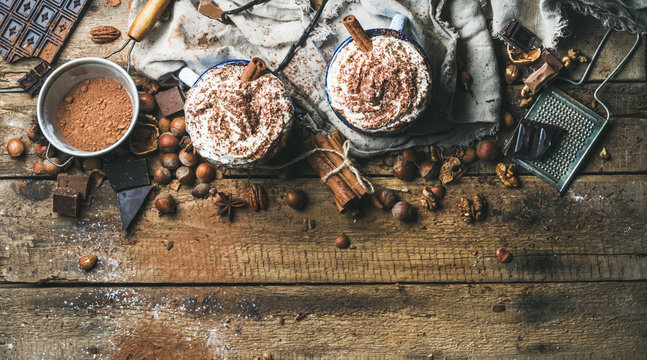 Hot chocolate with whipped cream and cinnamon sticks served with anise stars, different nuts and cocoa powder on rustic wooden background, top view, selective focus, copy space, horizontal composition