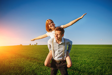 Smiling Man is holding on his back happy woman, who pulls out her arms and simulates a flight against the background of the blue sky and the green field