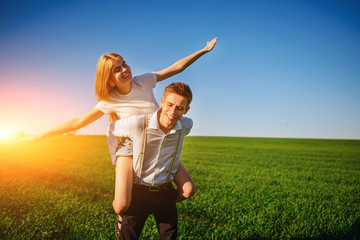 Smiling Man is holding on his back happy woman, who pulls out her arms and simulates a flight against the background of the blue sky and the green field