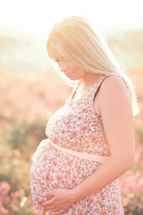 Fototapeta na wymiar Pregnant woman standing in sun light outdoors. Holding tummy. wearing stylish dress with floral pattern. Motherhood. Maternity.