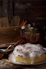 Glazed cinnabon buns on white craft plate with cup of coffee and cooper teapot on rustic wooden table opposite of wooden box with cinnamon sticks and wooden board
