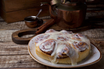 Glazed cinnabon buns on white craft plate with cup of coffee and cooper teapot on rustic wooden table opposite of wooden box with cinnamon sticks and wooden board