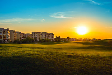 Fototapeta premium Green field for golf near apartment buildings, in dusk at sunset, tranquil place for rest