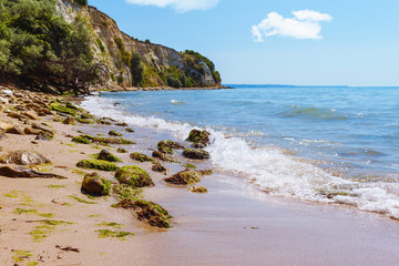 Black sea coast, seascape with stones on sand beach, cliffs above azure water, small wave splash, blue sky