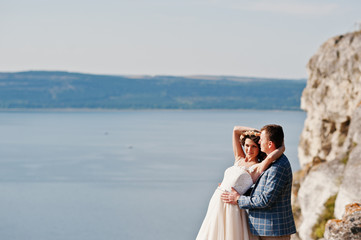 Fantastic wedding couple standing on the edge of rocky precipice with a perfect view of lake on the background.