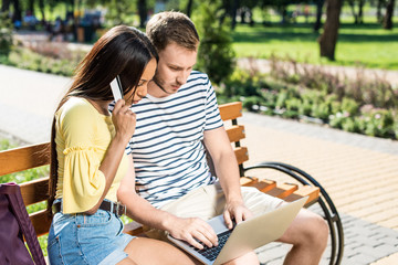 Fototapeta na wymiar Young multiethnic couple shopping online with laptop and credit card while sitting on bench in park