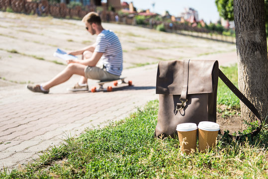 Close-up view of brown leather shoulder bag with disposable coffee cups and man sitting on skateboard behind
