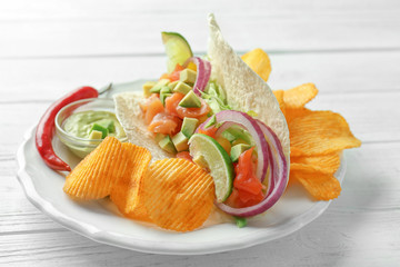 Tasty fish taco on plate with sauce and chips on white wooden background