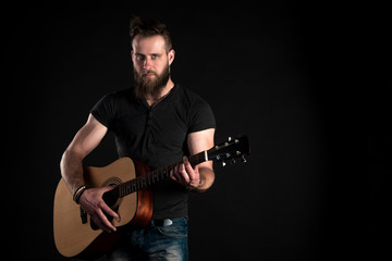 A charismatic and stylish man with a beard stands and plays an acoustic guitar on a black isolated background. Horizontal frame