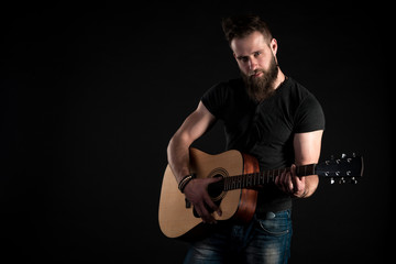 Obraz premium A charismatic and stylish man with a beard stands and plays an acoustic guitar on a black isolated background. Horizontal frame