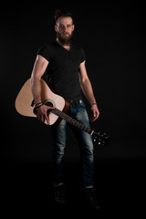 Fototapeta premium A charismatic and stylish man with a beard stands full-length with an acoustic guitar on a black isolated background. Vertical frame
