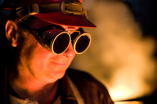 Portrait of the worker with protective goggles looking down, red color is a reflection of the molten metal. Very high heat and purple fringing. The inscription on the glasses is not a trademark.