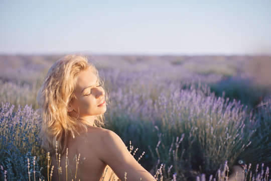 Beautiful blonde girl sit on the lavender field in golden sunset light. Romantic woman with Bare backt take sun bath on floral field having vacations in Provence, France.