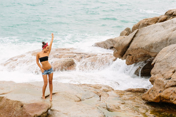 Pretty sporty lady in jeans short shorts, red headband and black bra stand on big stone on the beach during sea ocean storm. Big waves behind her. Concept of danger, sad, bad emotions.