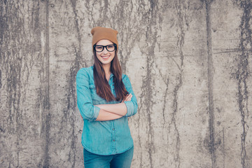 Attractive young girl student is standing on the concrete wall`s background outdoors, smiles, with crossed hands. She is wearing casual jeans outfit and brown hat, glasses
