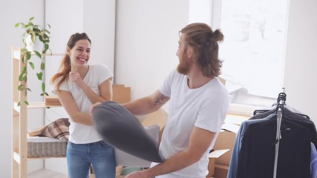Young Caucasian pair having fun and pillow fight in their new flat in slowmotion. Concept of happines and new life