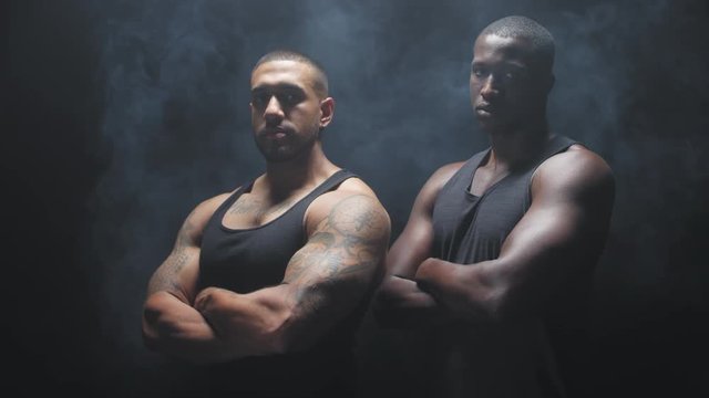 Portrait shot of two muscular black men staring at the camera with their arms crossed on a foggy dark background.
