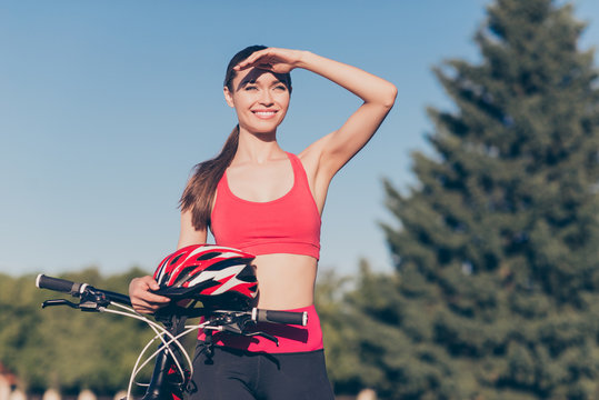 Great view! Cute young sporty lady is outside on a summer day, finished cicling on modern bike, in fashionable outfit, with helmet, looking far