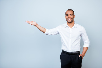 Young cheerful successful black lawyer on the pure light blue background is smiling, standing in formal wear and is holding the copyspace