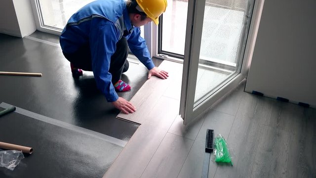 Skilled handyman guy with yellow hard hat installing laminate boards on floor