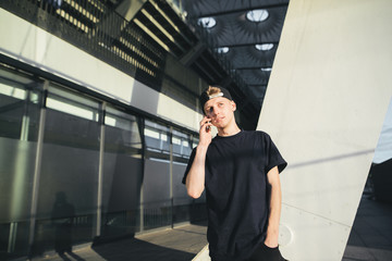 A stylish young teenager in a black T-shirt and cap is talking on the phone with the background of modern architecture.