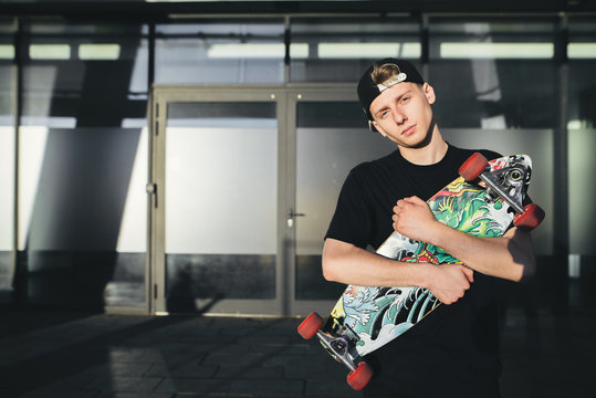 Portrait of a young beautiful skateboard with a painted board in his hands against the background of a wall of modern architecture. A teenager with a longboard in his hands poses after a ride.