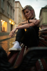 Young woman sits on wet bench on city street in evening in rain.