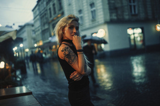 Young woman with tattoo on shoulder stands on city street in evening in rain.