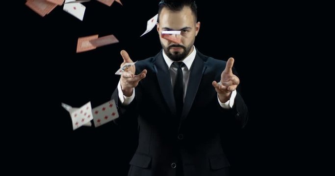Professional Young Magician in a Dark Suit Throws Two Jets of Cards into the Air in Slow Motion. Background is Black. Shot on RED EPIC-W 8K Helium Cinema Camera.