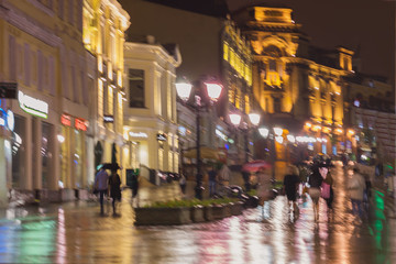 Abstract blurred background of people under umbrella walk city street in rain. Bright illumination, reflection in wet pavement from shop windows, street lamps. Lifestyle of modern city, seasons