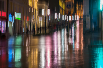Abstract bright blurred background with unidentified people city street in rainy night. Vivid illumination, reflection in wet pavement from shop windows, street lamps. Concept active lifestyle