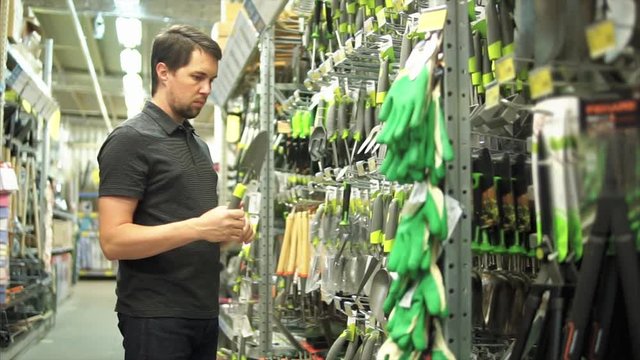 Man gardener in the hardware shop choosing small gardener shovel for his garden. Shelves of garden tools on background