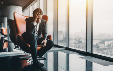 Businessman in formal suit is sitting on orange armchair and holding his head in depression after...