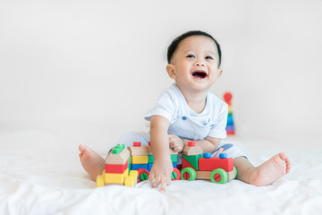 Adorable Asian baby boy 9 months sitting on bed and playing with color wooden train toys at home..