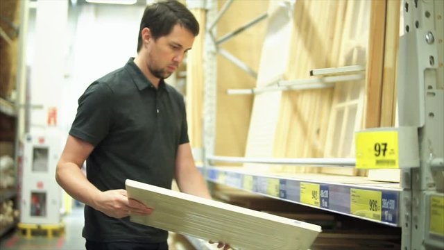 Male customer in the hardware store standing near shelf of wooden crafts. He taking the wooden plank and knocking on it.
