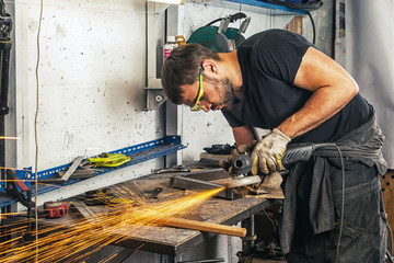 A young man welder in a black T-shirt, glasses and construction gloves  grinder metal an angle...