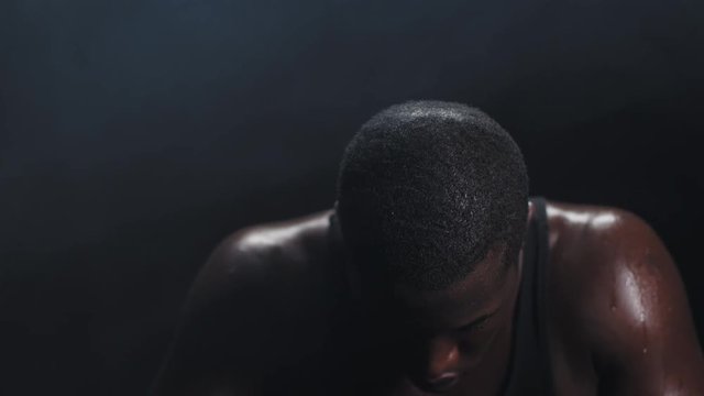 Zooming shot of a muscular black man wiping the sweat off his forehead on a foggy dark background