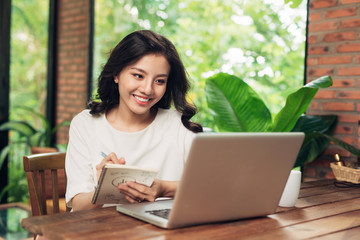 Young smiling asian woman sitting at table in cafe and writing in notebook