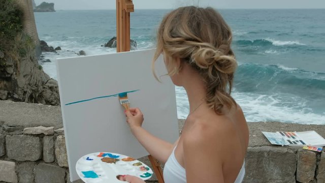 Young woman paints picture standing by seaside on summer day. Located at parapet against backdrop of raging waters of Mediterranean Sea, she holds palette with watercolor and neat strokes apply blue