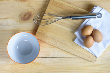 Eggs with whisk and empty ornage bowl on wooden plate