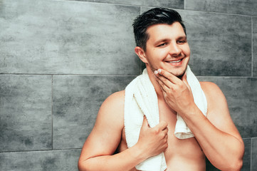Handsome young man staying in bathroom with white towel on shoulders. Brunette muscle guy with shirtless torso holding towel smile and touching his face, checking his beard Wellness skincare concept.
