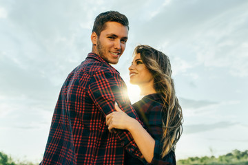 Awesome couple in a field sunset background. Handsome blond guy with a beard in plaid shirt gentle hugs a girl with blond hair in plaid shirt looking in his eyes. Lifestyle and travel concept.