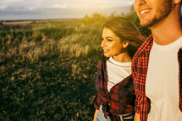 Cropped handsome guy with a beard in a plaid shirt hugs and holds the hand of a beautiful girl in a plaid shirt in a field at sunset. Stylish happy couple smiling on dating. Lifestyle and travel.